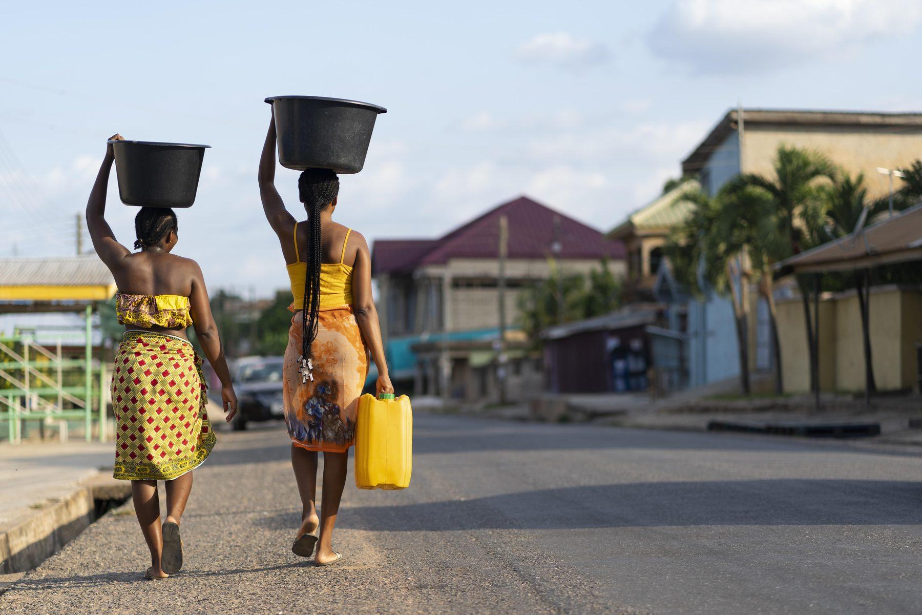 Two women walking home along a road at the end of the day, basins carried on their heads and a jerrican in hand.
