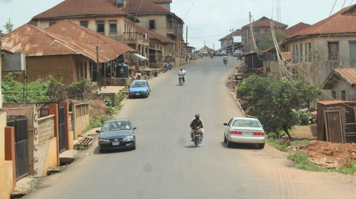 A street through Ilobu: rust-red roofs on both sides, parked cars along the verge, a rider coming up the middle of the road.